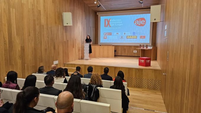 Evento en una sala con un presentador y audiencia atenta, fondo de pantallas con logotipos.