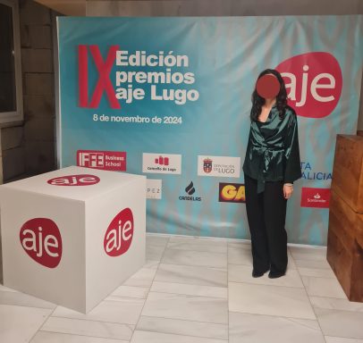 Mujer de pie junto a un panel de fondo de los IX Premios AJE Lugo.